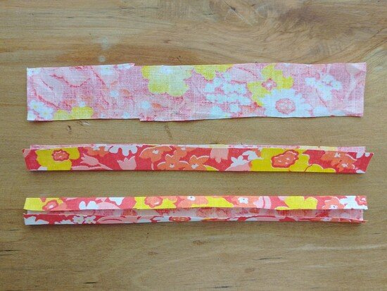 Three long strips of floral fabric laid out flat on a wooden surface. From the top a strip, below this, a strip folded lengthewise in half, and at the bottom the strip folded in half again towards the center crease