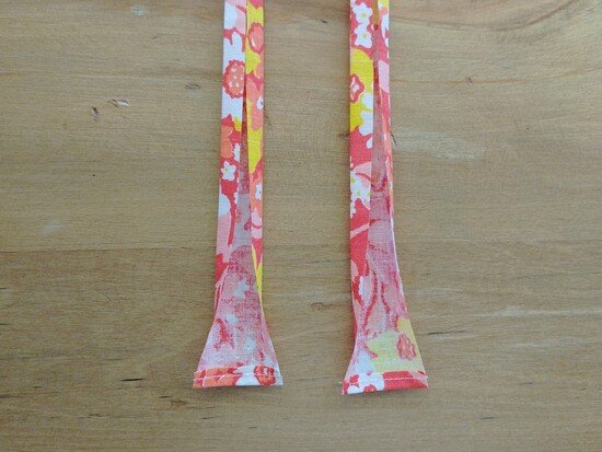 Two narrow floral fabric single fold binding strips with the bottom edges folded over and stitched laid flat on a wooden surface.