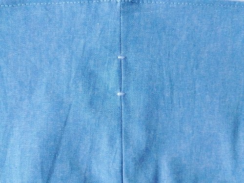 A close-up of blue chambray fabric showing two white bar tacks stitched above and below a small opening in the seam.