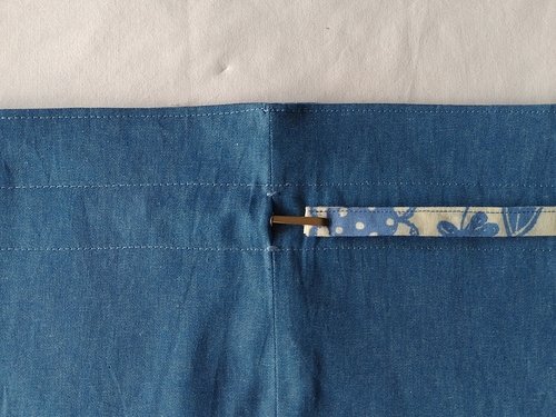 A metal bodkin and floral drawstring being threaded through a small opening in a blue chambray seam, located between two horizontal rows of stitching.