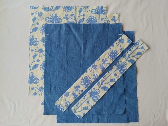 Four rectangles of fabric,two floral and two solid blue chambray,stacked with two floral fabric strips placed diagonally on top.