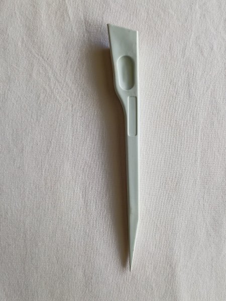 A slim, light gray finger pressing tool for sewing, positioned vertically on a white fabric background. The tool has a wide, flat top with a thumb indent and tapers down to a sharp point at the bottom.