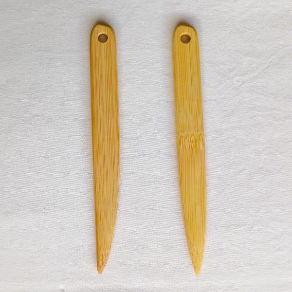 Two long, bamboo wooden point turner tools are positioned vertically side-by-side on a white fabric background. Both tools have a rounded top with a small circular hole for hanging, and they taper down.