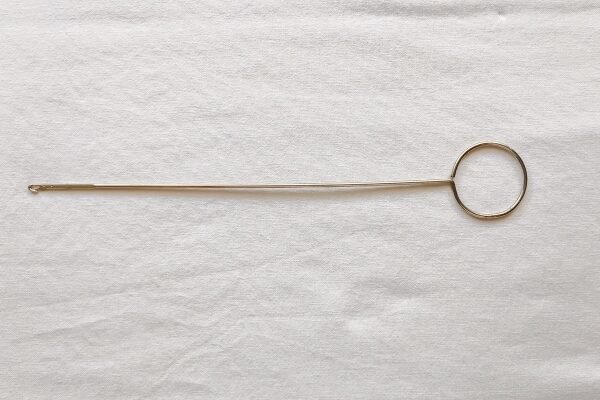 A long, thin metal loop turner tool lies horizontally against a white fabric background. It features a large circular ring handle on the right and a tiny latch hook at the tip of the slender rod on the left.