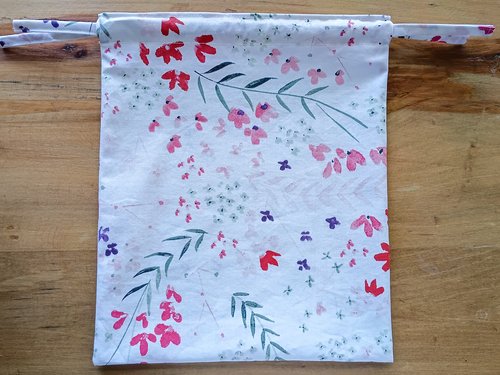 A floral fabric bag laid flat on a wooden surface, with the top gathered slightly and two fabric drawstrings extending from the sides.