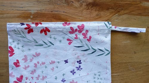 A white floral drawstring bag laying flat on a wooden surface with a fabric loop on the right side.