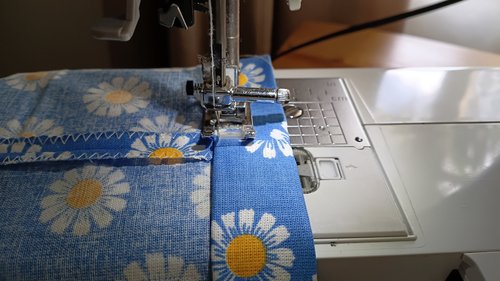 A sewing machine stitching down the folded top edge of a blue daisy fabric.