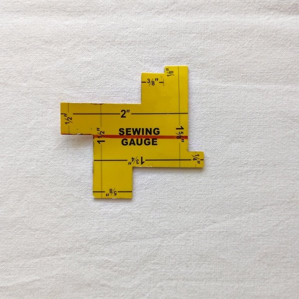 A small, yellow, plastic sewing gauge with various measurements marked in black and red, shown against a white fabric background. The tool features several notched edges labeled with common sewing increments like 5/8, 1/4, and 2 inches.