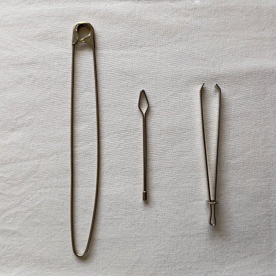 Three different metal bodkin tools for sewing are arranged vertically on a white fabric background. From left to right: a long safety-pin style bodkin, a small needle-like bodkin with a large diamond-shaped eye, and a tweezer-style bodkin with a sliding ring to lock the g
