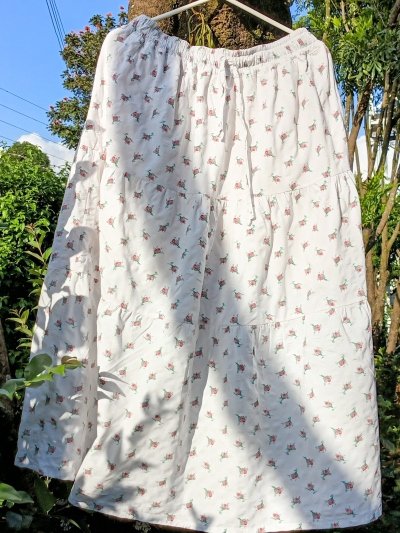 A white, three-tiered maxi skirt with a small red floral print hanging on a hanger outdoors.