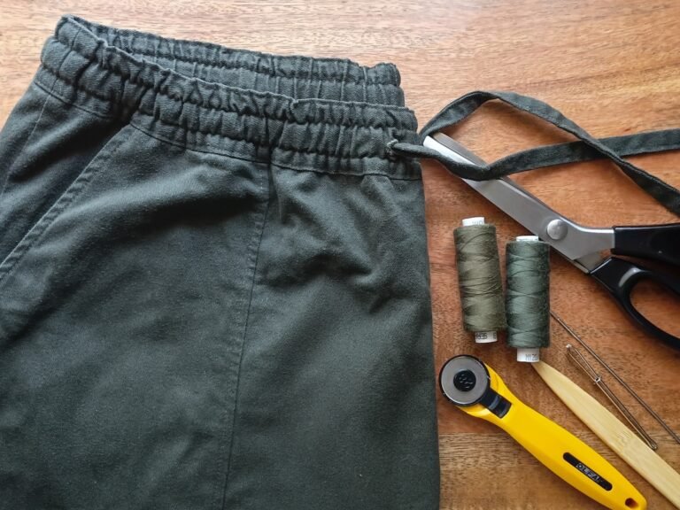 Dark green elastic-waist pants with sewing tools and thread on a wooden table.