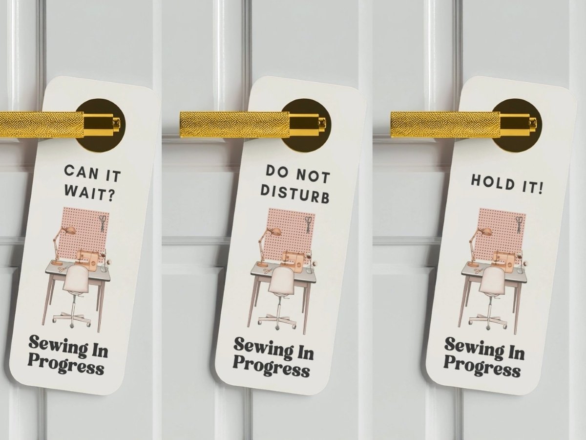 Three white door hangers with gold handles, each showing a sewing workspace illustration and the words “Sewing In Progress,” with different messages at the top: “Can It Wait?”, “Do Not Disturb,” and “Hold It!”