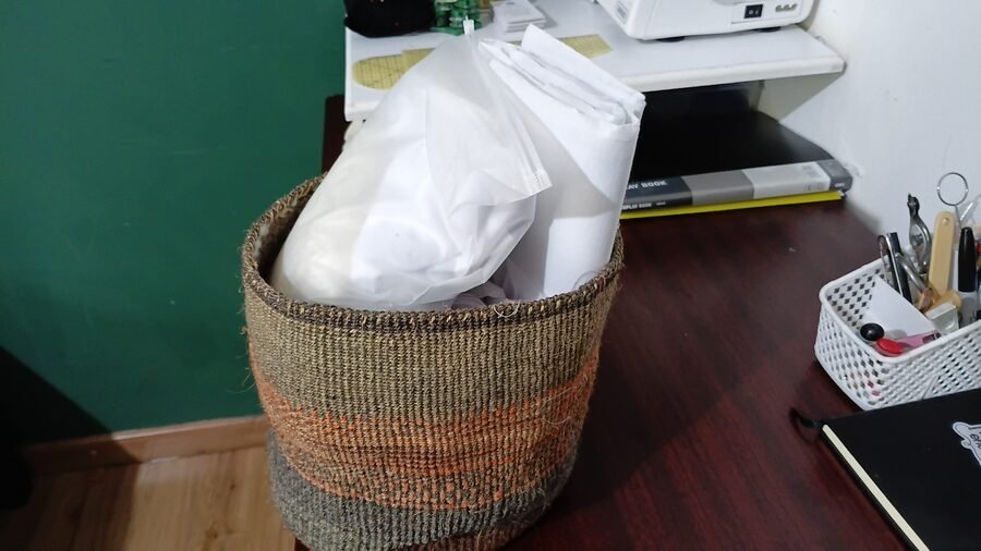 Woven basket holding rolls of interfacing, stabilizers, and other sewing materials beside the sewing workstation.