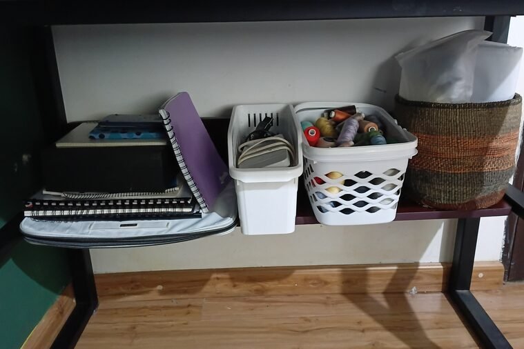 Under-table storage at a sewing workstation, with notebooks, sewing patterns, machine accessories, thread, and baskets containing sewing supplies and interfacing.