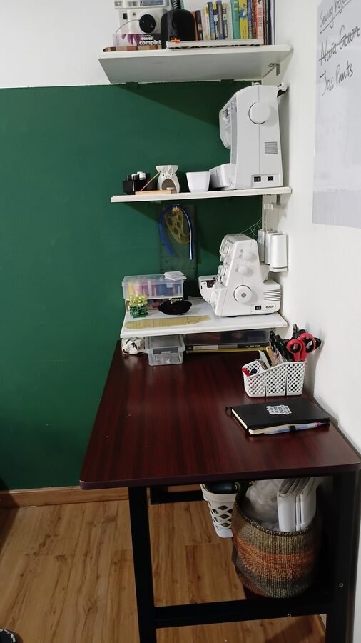 Sewing corner with a dark wood desk, sewing machine and serger on wall-mounted shelves, and sewing supplies stored in bins and baskets.