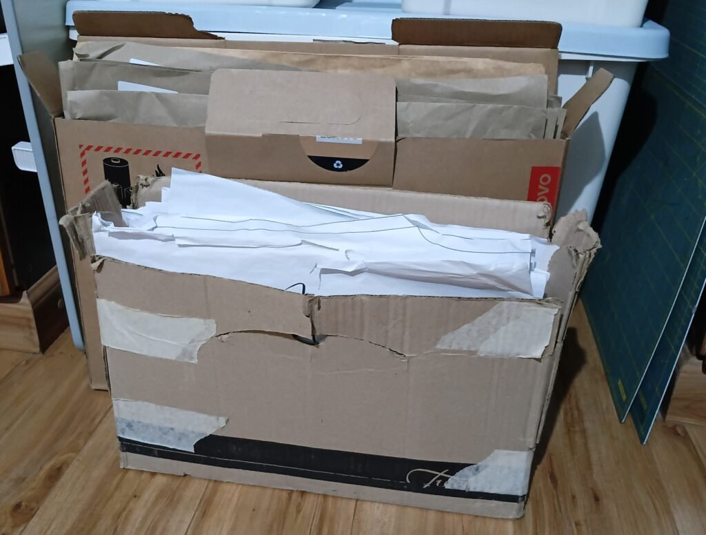 Cardboard boxes filled with sewing patterns stored upright and organized by envelope, with a cutting mat visible nearby.