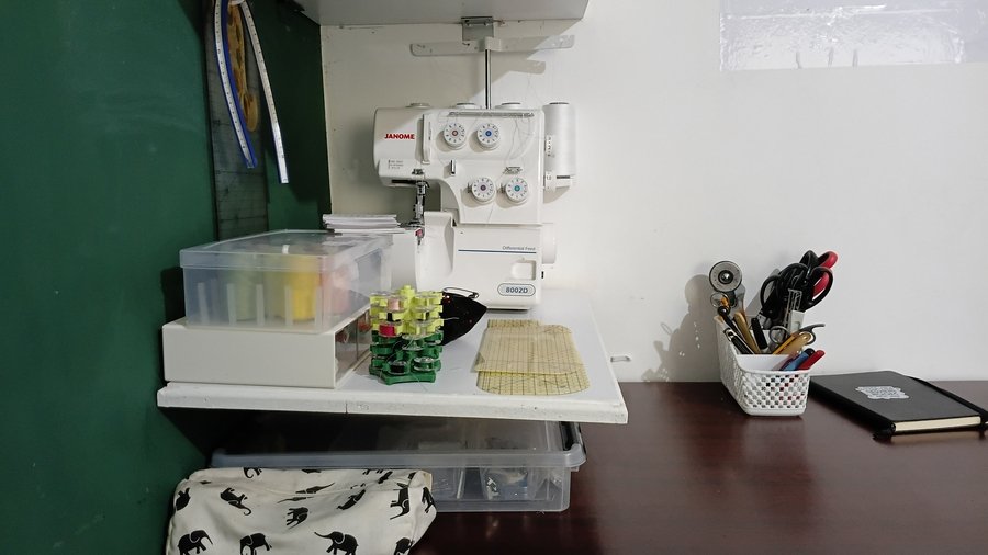 Close-up of a serger and sewing supplies on a wall-mounted shelf above a worktable, with thread spools, storage containers, cutting tools, and a notebook nearby.