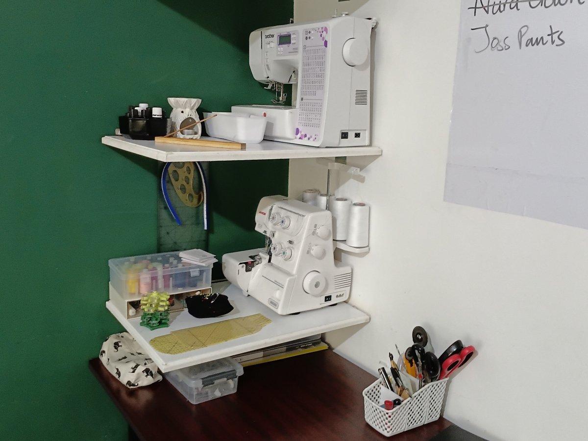 Compact sewing station in a bedroom corner, featuring wall-mounted shelves with a sewing machine, serger, organized sewing supplies, and a small worktable.