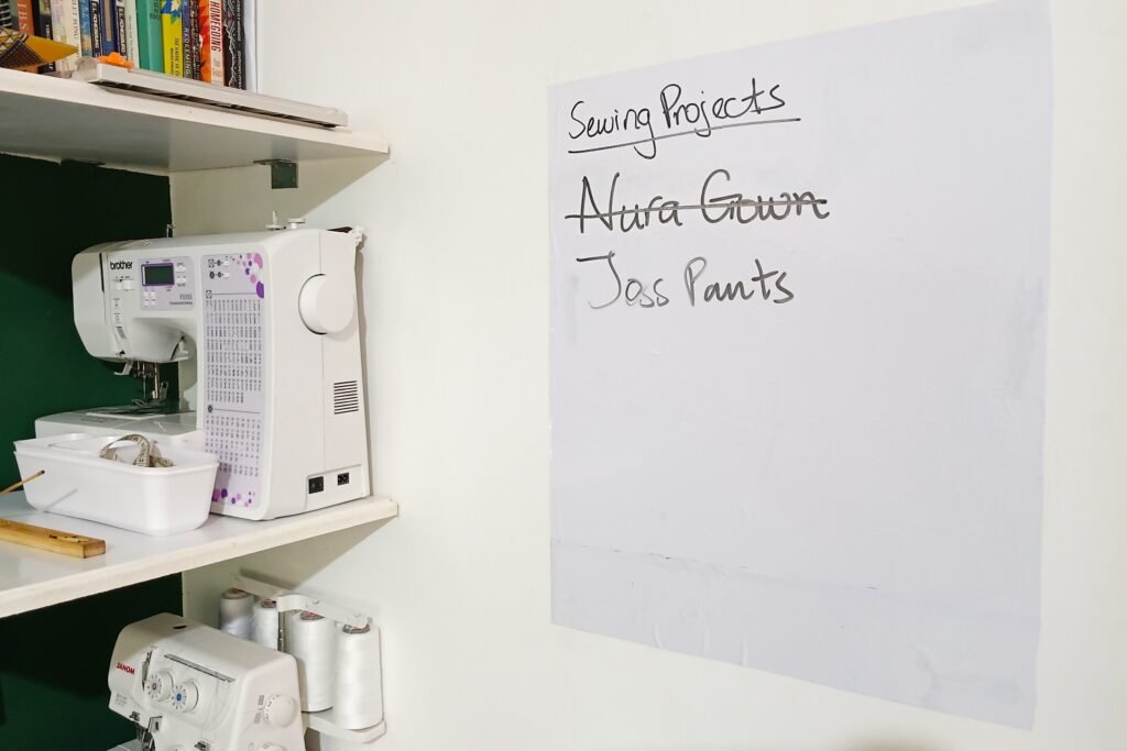 Sewing workstation with a sewing machine on wall-mounted shelves beside a handwritten project board listing current sewing projects.