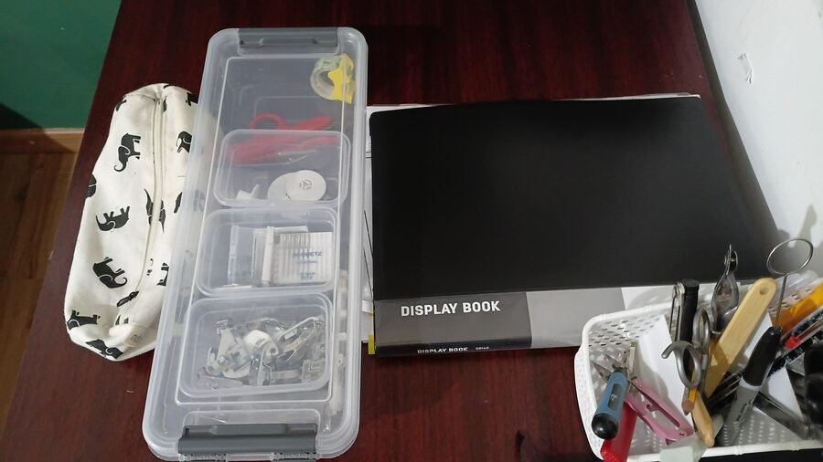 Sewing tools and supplies organized on a worktable, including a storage box of machine accessories, a document binder, and a container of frequently used tools.