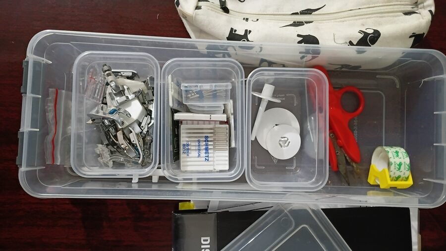 Close-up of a sewing accessory storage box containing presser feet, machine needles, bobbins, scissors, and other sewing essentials.
