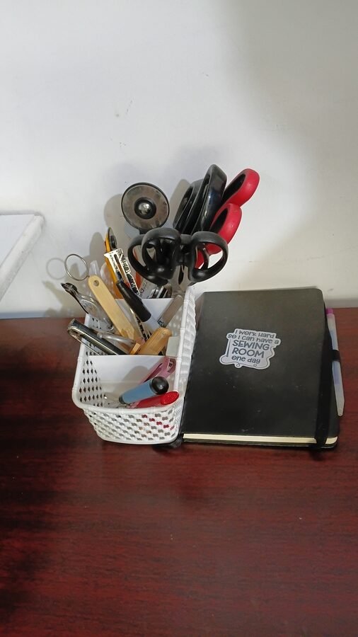 Sewing tools stored in a small desktop organizer beside a notebook on a sewing worktable.