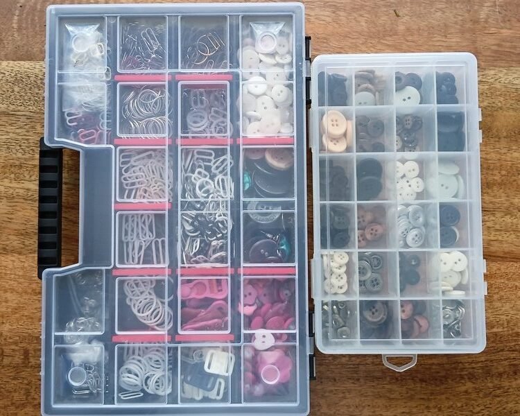Two clear organizer boxes of buttons, snaps, and bra hardware sitting on a wooden table.