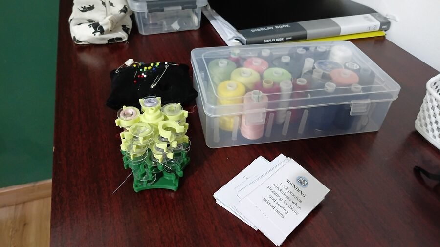 Sewing supplies arranged on a worktable, including a thread storage box, bobbin holder, pin cushion, and a stack of sewing affirmation cards.