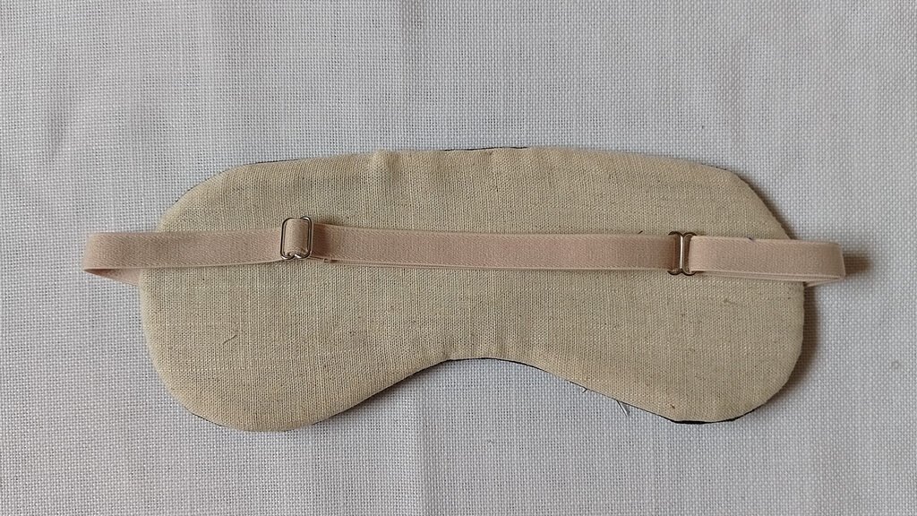 The back of a sleep mask. It has beige linen fabric and a beige adjustable strap