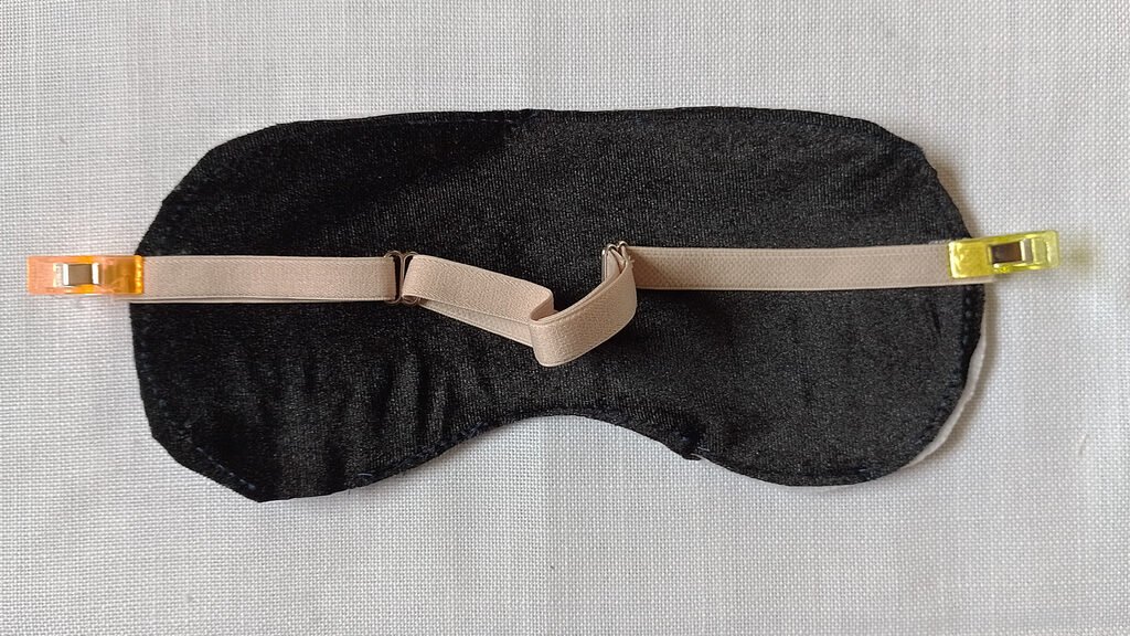 A beige adjustable strap pinned onto a mask front. the mask front is a piece of black velvet and batting basted together.