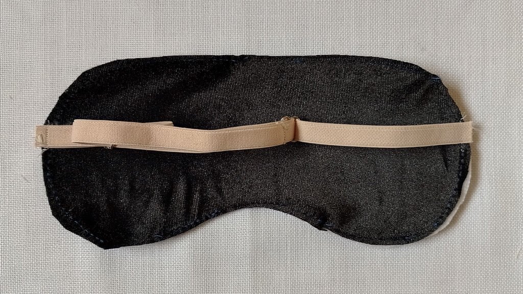 The front a sleep mask made from black velvet with a beige adjustable strap sewn down at the edges