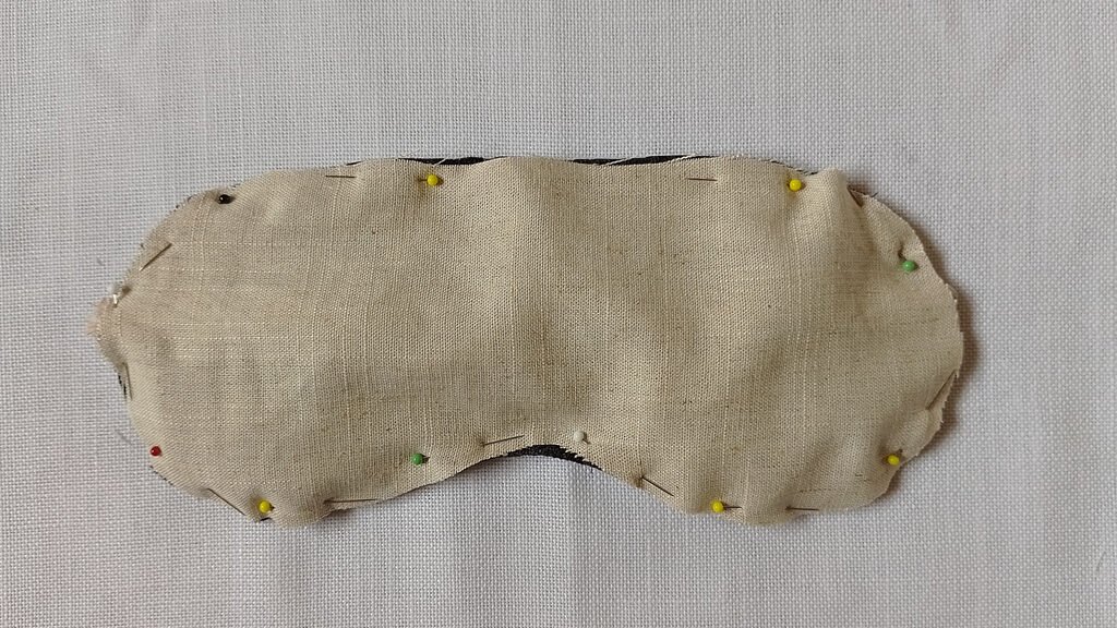 A sleep mask being constructed. The back piece, a beige linen, is pined on top of the rest of the mask.