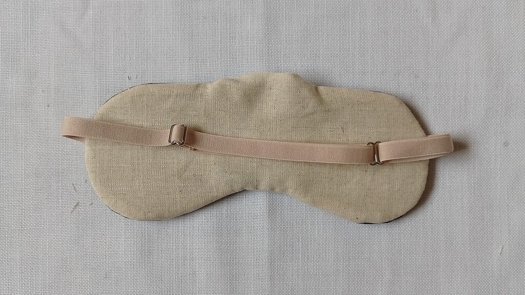 The back of a sleep mask with a gaping whole at the top. It has beige linen fabric and a beige adjustable strap.