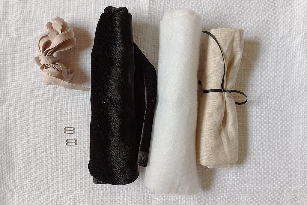 The supplies needed for the sleep mask sewing project which in this photo include a beige linen offcut, a black velvet offcut, beige bra strap elastic, two silver sliders and batting