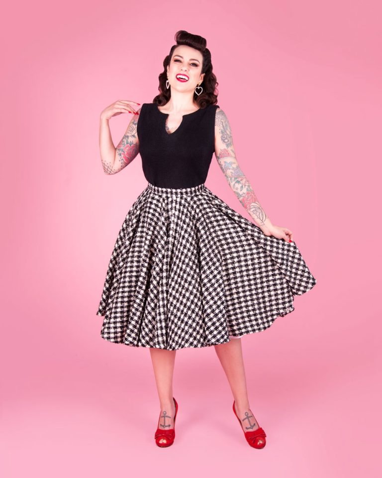 Woman in a black sleeveless top and black and white check circle skirt with red peep-toe heels on a pink background