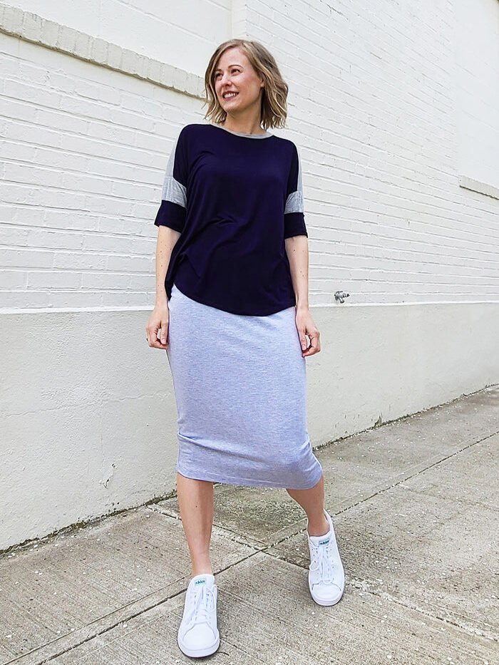 Woman in a navy colorblock top and gray knit pencil skirt with white sneakers, standing against a brick wall