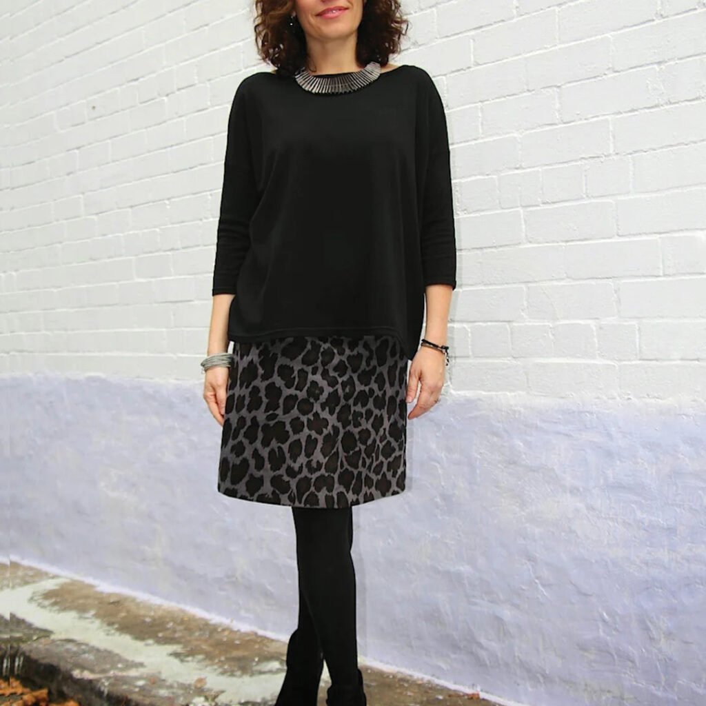 Woman in a black oversized top and gray leopard print A-line skirt with black tights and boots, standing against a brick wall, Libby Skirt free pattern by Tessuti