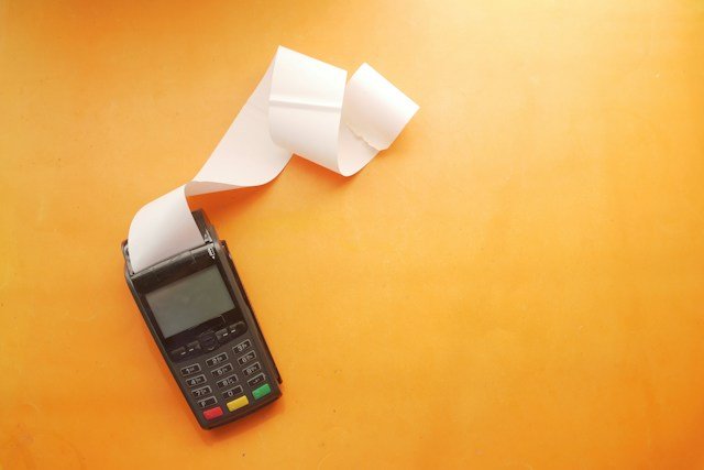 A payment terminal with a roll of white receipt paper curling out onto an orange surface.