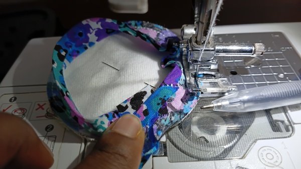 Hand guiding a flannel round with floral bias tape under the sewing machine needle, with a pen resting beside it
