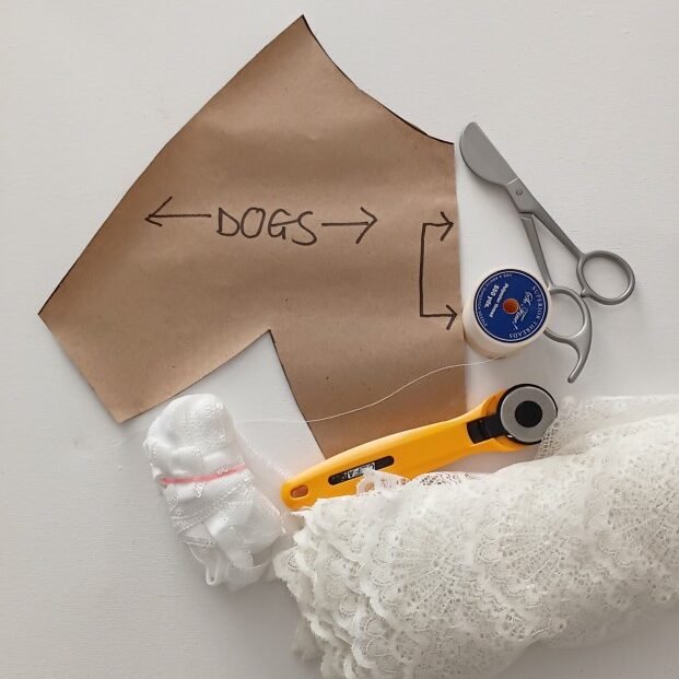 Flat lay photograph of bralette sewing supplies on a white surface, including a brown paper pattern piece with handwritten markings, fabric scissors, a yellow rotary cutter, a blue measuring tape reel, and white lace fabric with elastic trim.